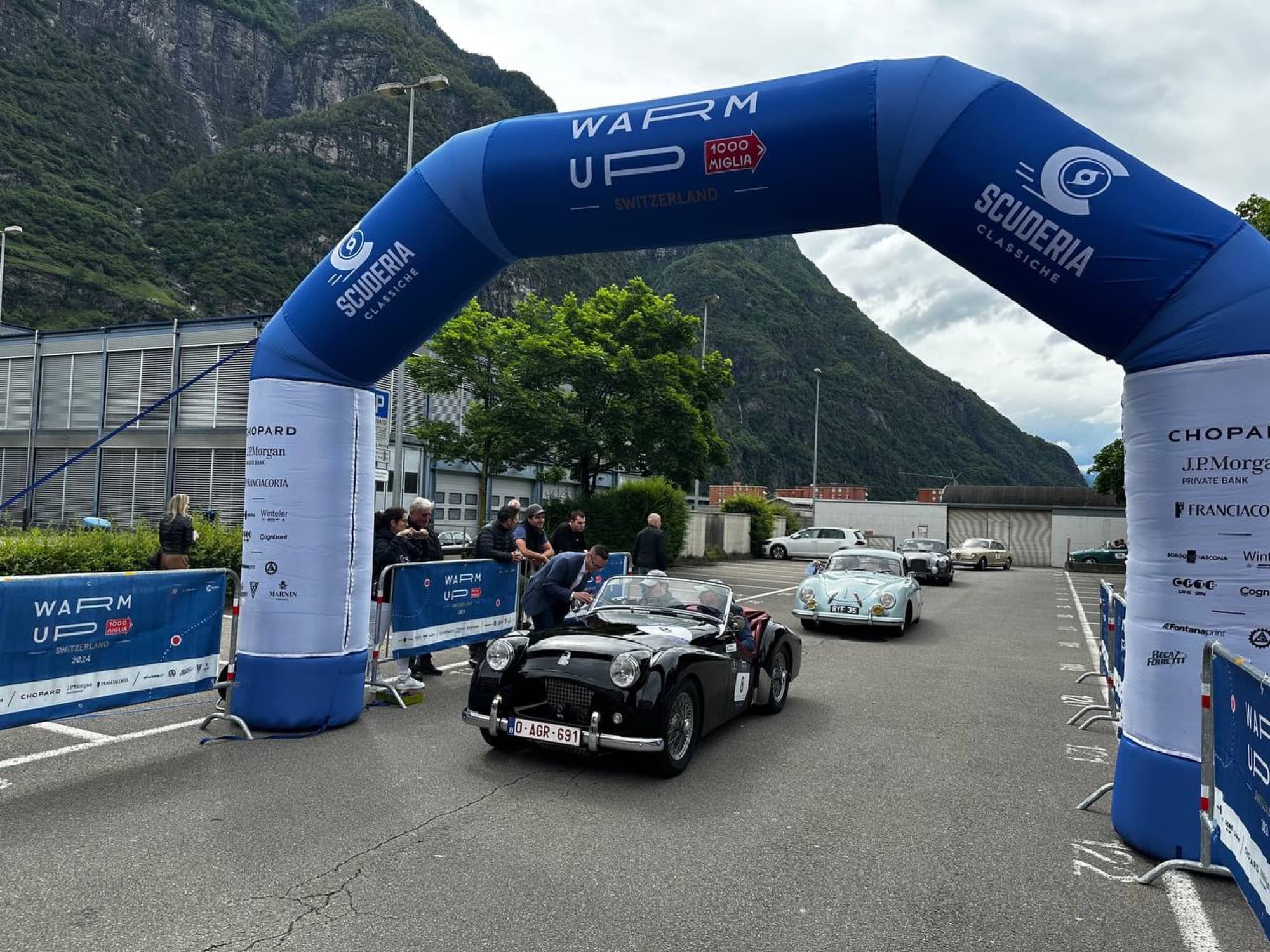 Arco blu WARM UP · 1000 MIGLIA con il logo Scuderia Classiche, auto d'epoca in transito, montagne sullo sfondo.